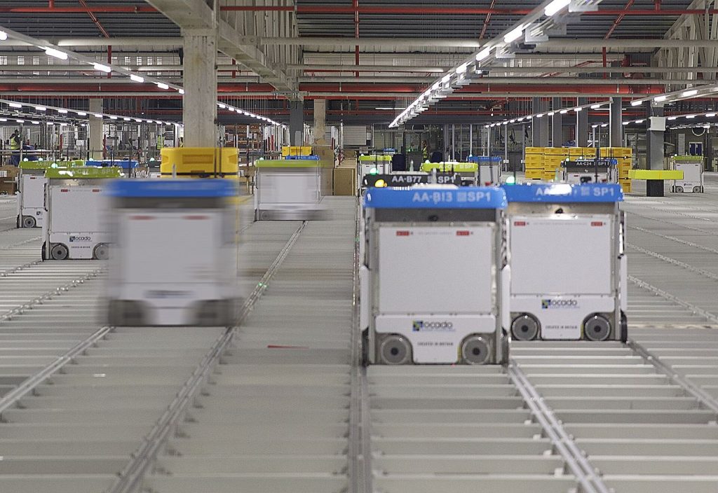 Orange warehouse robots carrying tall shelving units across a busy fulfilment centre floor.